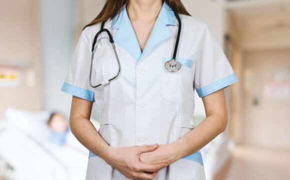 Healthcare professional in a white coat with a stethoscope, standing with hands clasped